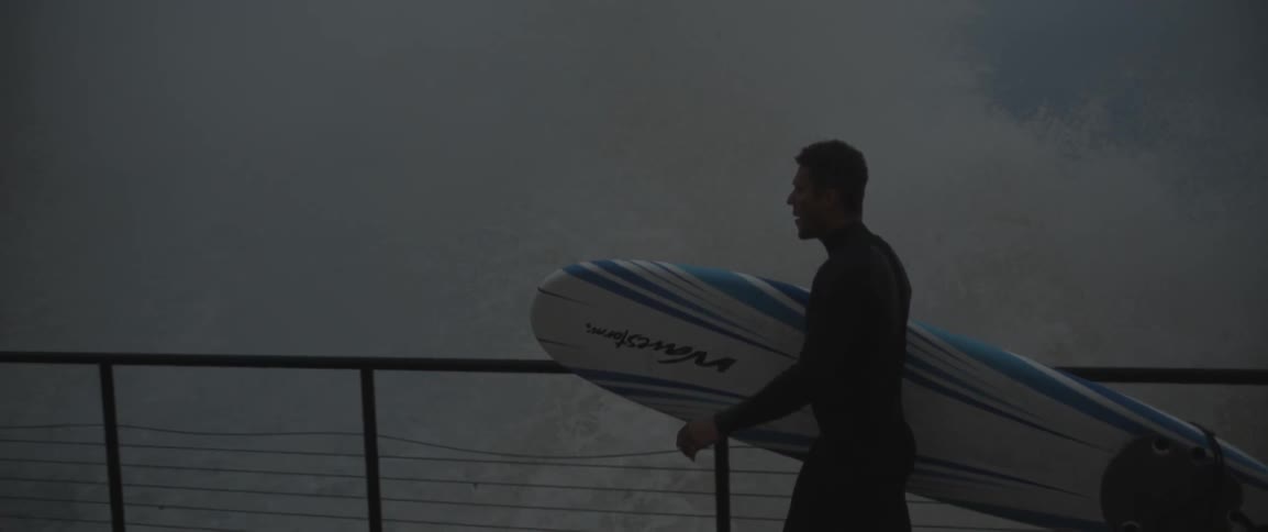 Man holding surfboard walking on walkway near crashing waves