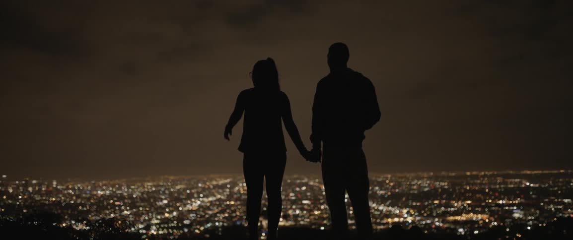 A man and woman hold hands while overlooking city lights at night