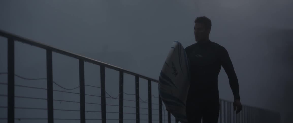 A man walks along the fence carrying a surfboard as waves hit the fence
