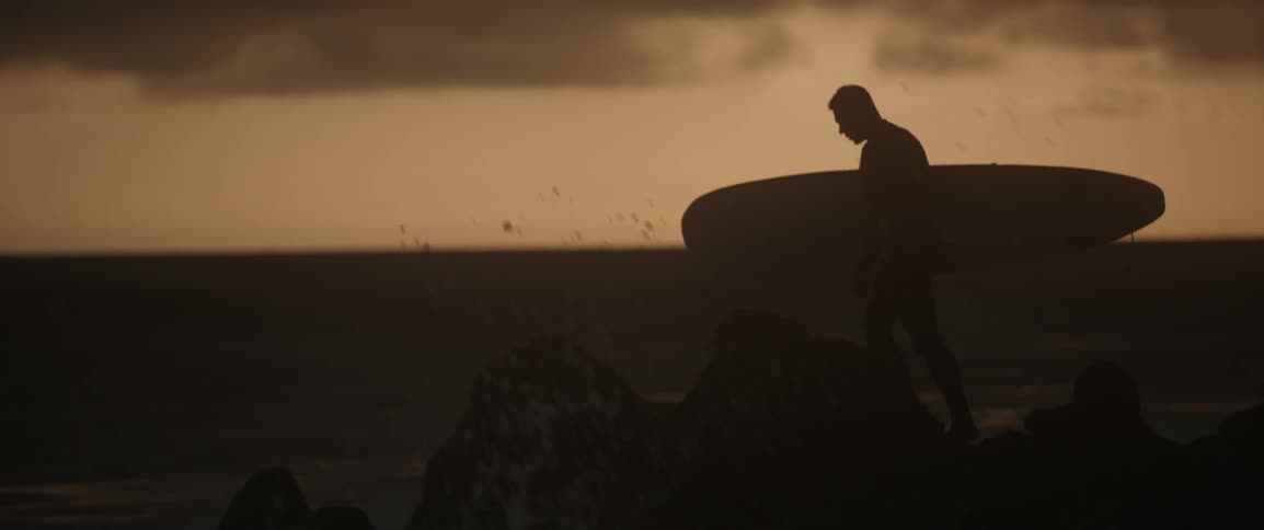 A man walks carrying a surfboard at dawn