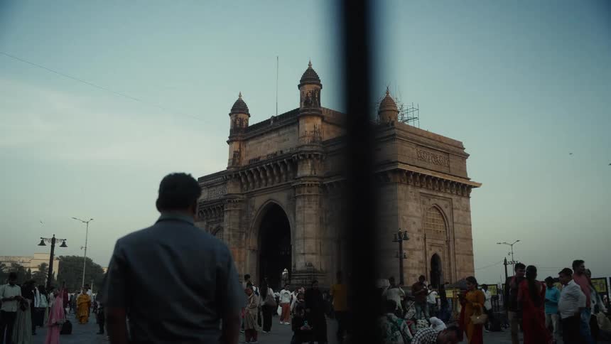 A large stone archway monument is seen from behind a foreground persons shoulder with a large crowd of people gathering and moving around the base of the historic structure