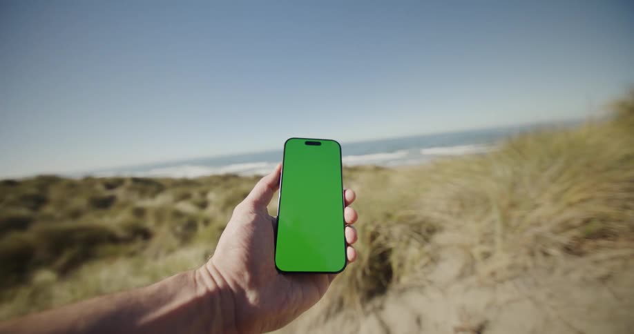 Hand holds phone in front of dunes and sea under clear sky
