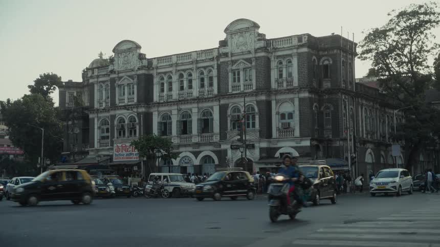 A large multistory historic building located at a busy city intersection where various automobiles taxis and motorcycles move through the frame as a crowd of people gathers near the buildings ground level