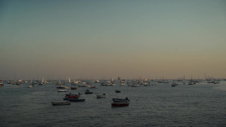 A wide static shot shows a vast expanse of dark water where dozens of various sizes of sailboats and small fishing vessels are anchored set against a muted gradient sky during the transition of dusk