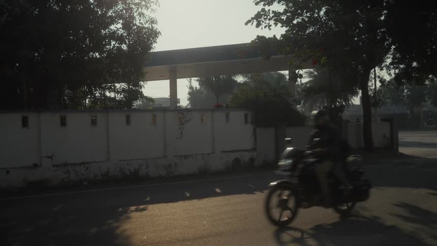 This very short clip is filmed from the roadside showing a gas station canopy visible over a low lightcolored walla person on a motorbike quickly drives past the camera from right to left in the foreground