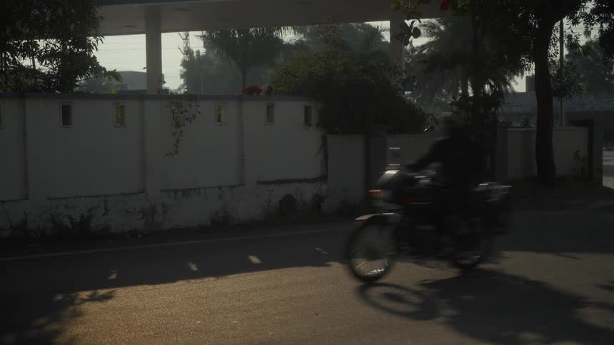 The video shows a quick shot of a street scene dominated by a low whitewashed boundary wall and shadowed trees behind it a dark blurred figure on a motorcycle quickly moves from left to right across the foreground