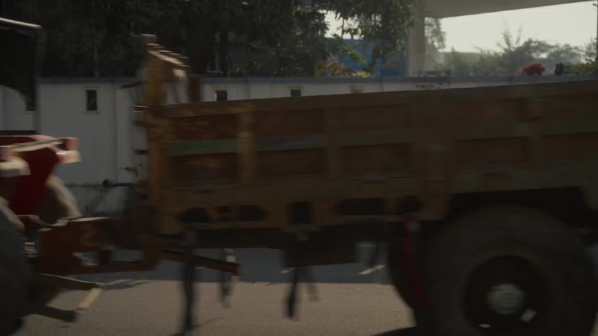 Red tractor with canopy pulls metal trailer in motion along paved road beside greenery