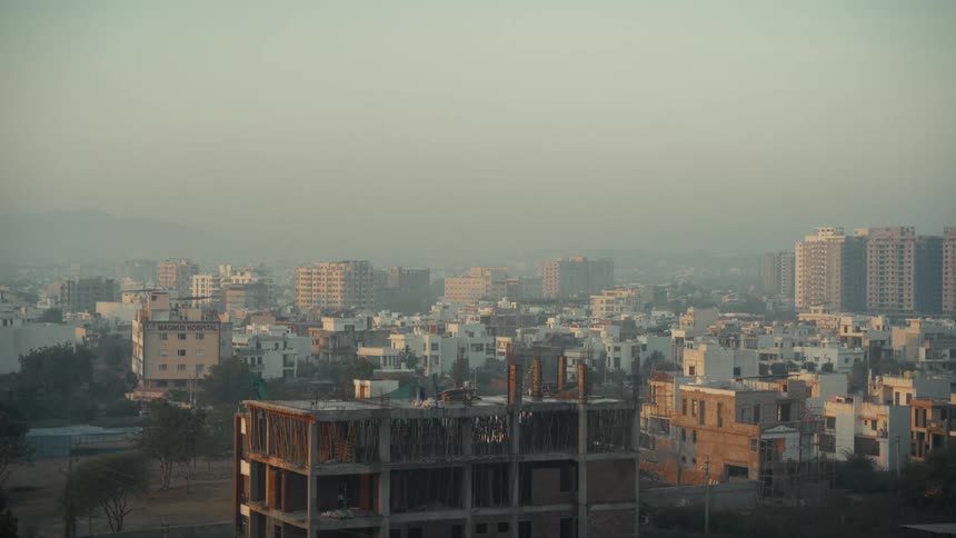 This is a wide elevated view of a dense sprawling cityscape under a hazy or smoggy skyin the foreground an unfinished building under construction contrasts with completed residential and commercial structures stretching toward distant hills