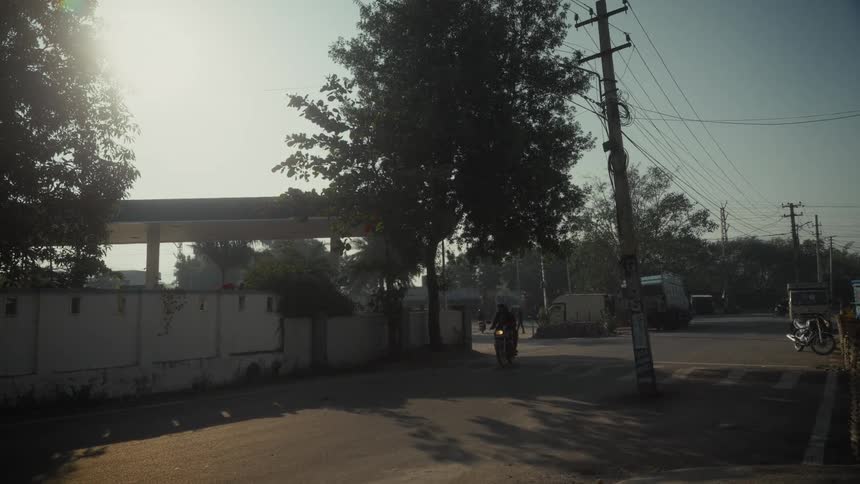 Motorcyclist moves along urban road under high sun amid vehicles poles and roadside greenery