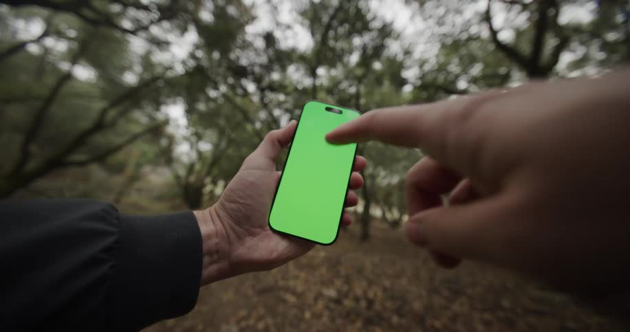 The video features a closeup firstperson shot of a person holding a modern smartphone horizontally in a forest settingthe phones screen is a solid green color for easy visual effects replacement and a second hand briefly enters the frame to inter
