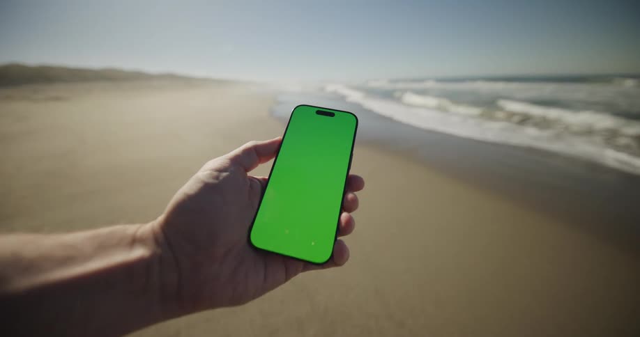 Man holds a mobile phone with a glowing green screen as he walks on the beach and waves roll in to shore