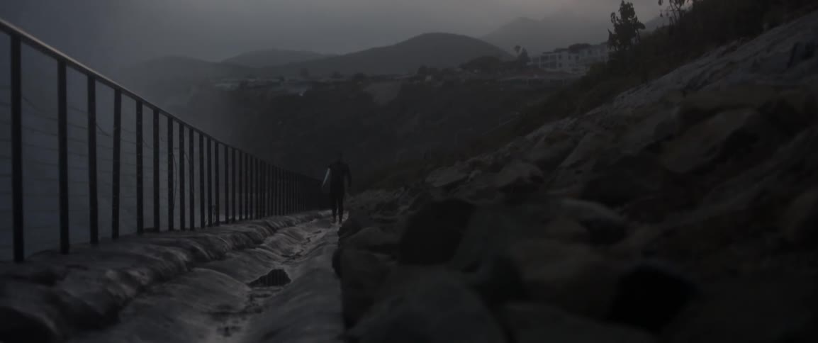 A man walks with a surfboard by a fence near the ocean