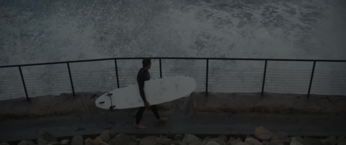 Man holding surfboard walking on walkway near crashing waves