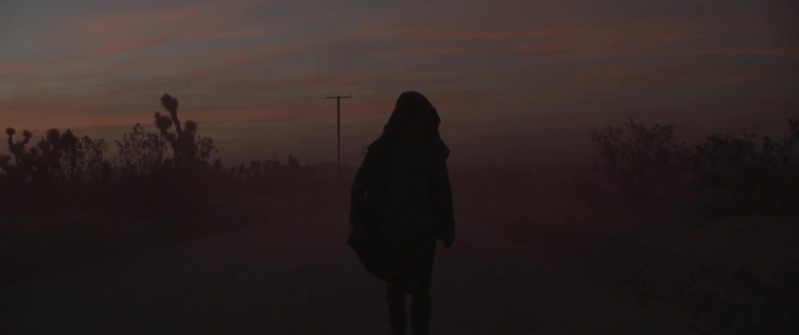 A hooded figured walks on a foggy road at sunset