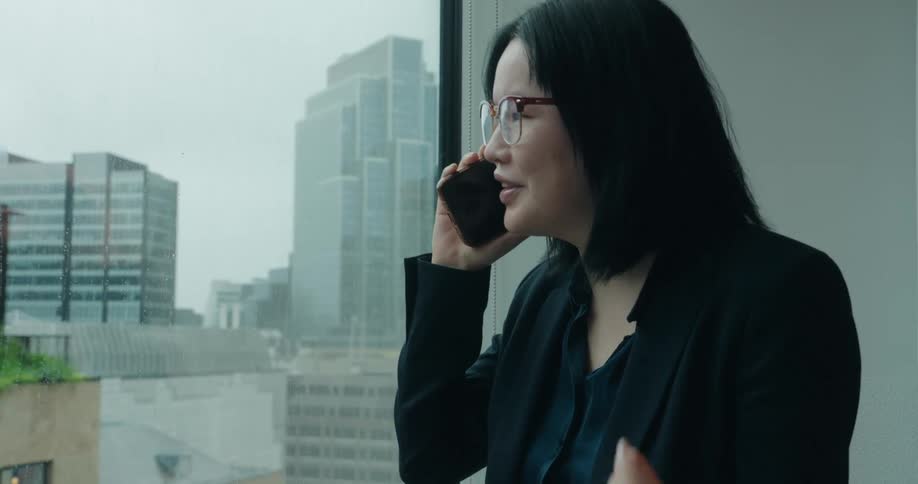 A middleaged asian woman dressed in a black suit and wearing glasses stands beside a glass window in a highrise office at midday talking on the phone