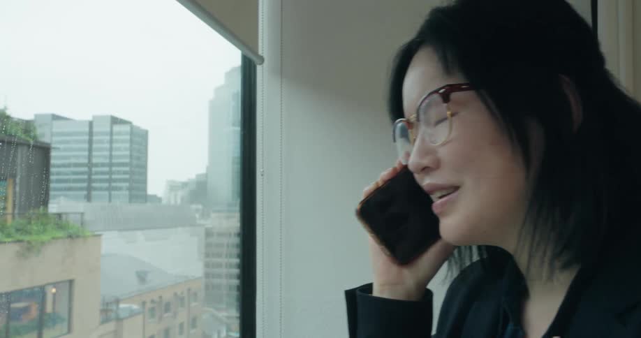 Young asian woman talking on the phone next to a window showing buildings on a metropolitan area outdoors during day time