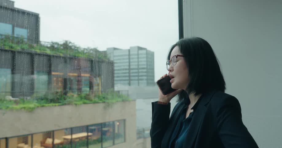 Woman talks on the phone leaning against a window on a rainy day