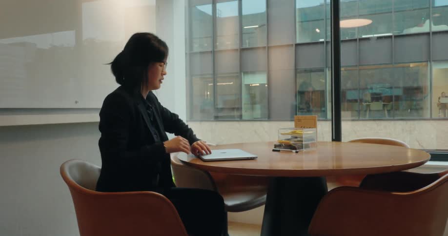 A woman in a black business suit walks into a glass walled meeting room sits down at a round table and opens her laptop to start working