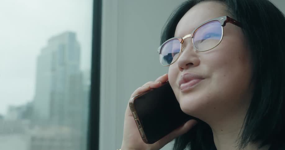 Woman standing near a window with a view of downtown talks into a smartphone during the daytime