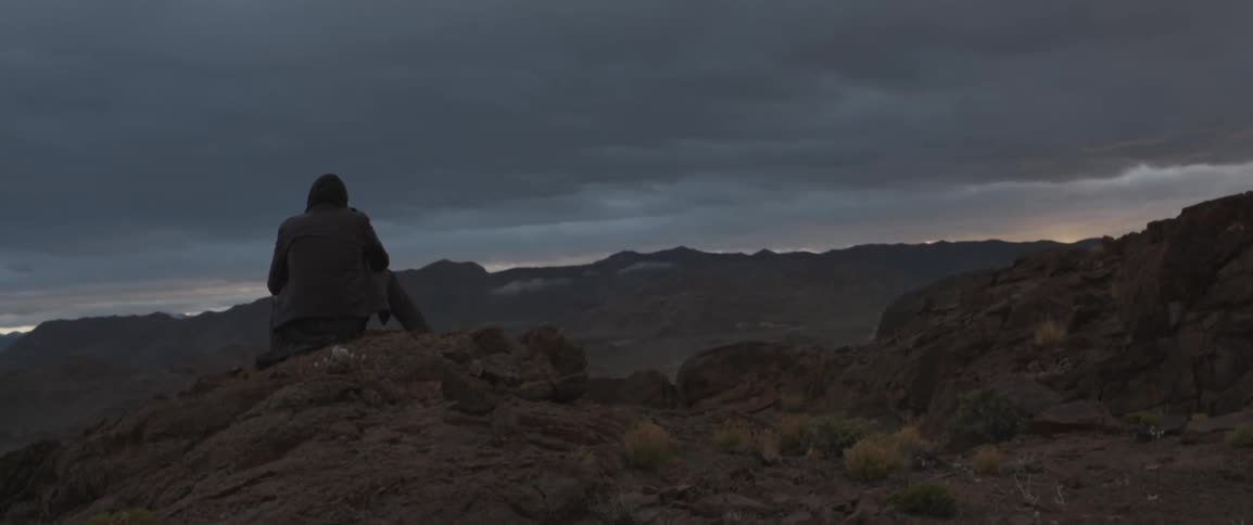 Man sits atop a rocky cliff looking out at mountains