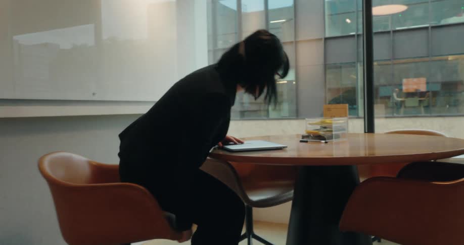 Female office worker sits in a meeting room with a view of another office building and types on a laptop