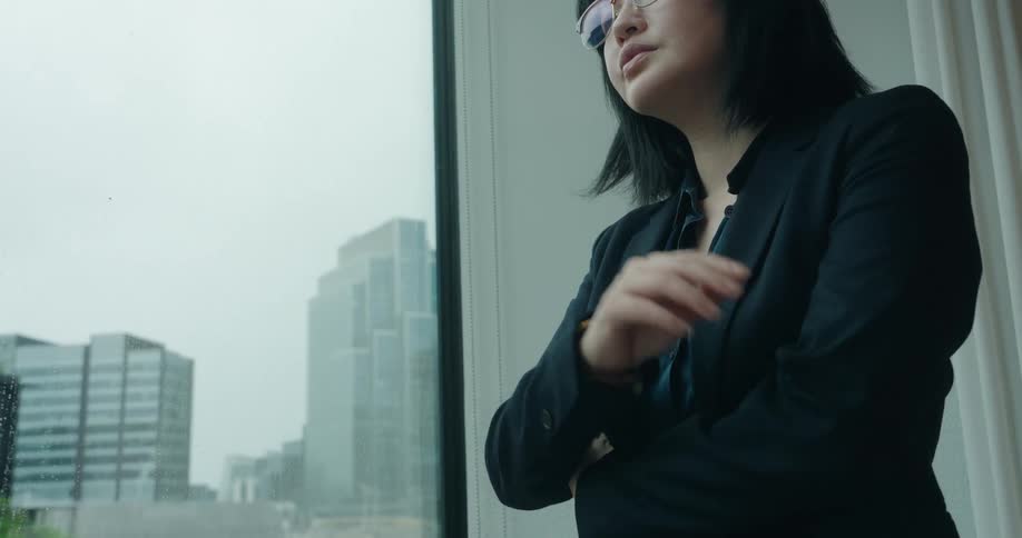 A contemplative professional woman in a business suit stands indoors by a large window