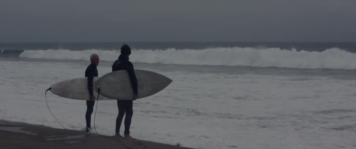 Two people stand with surfboards by the shore