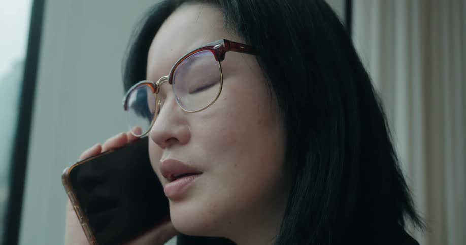A young asian woman wearing glasses stands beside a glass window inside an office building answering a phone call
