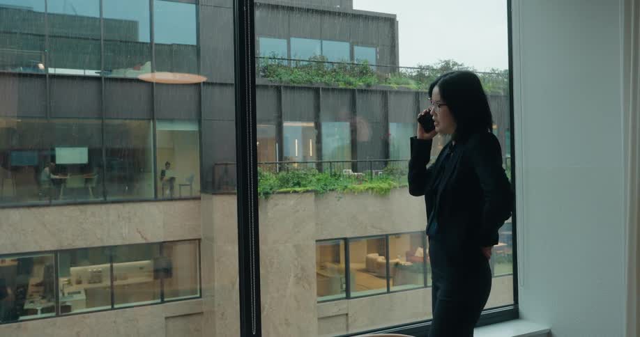A woman with short black hair wearing a black business suit stands facing a large window holding a mobile phone to her ear and speaking while looking out at an adjacent building on a overcast day