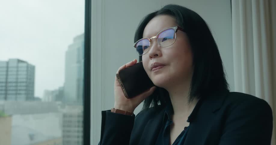 Woman stands next to a window with a view of downtown buildings as she talks on her phone