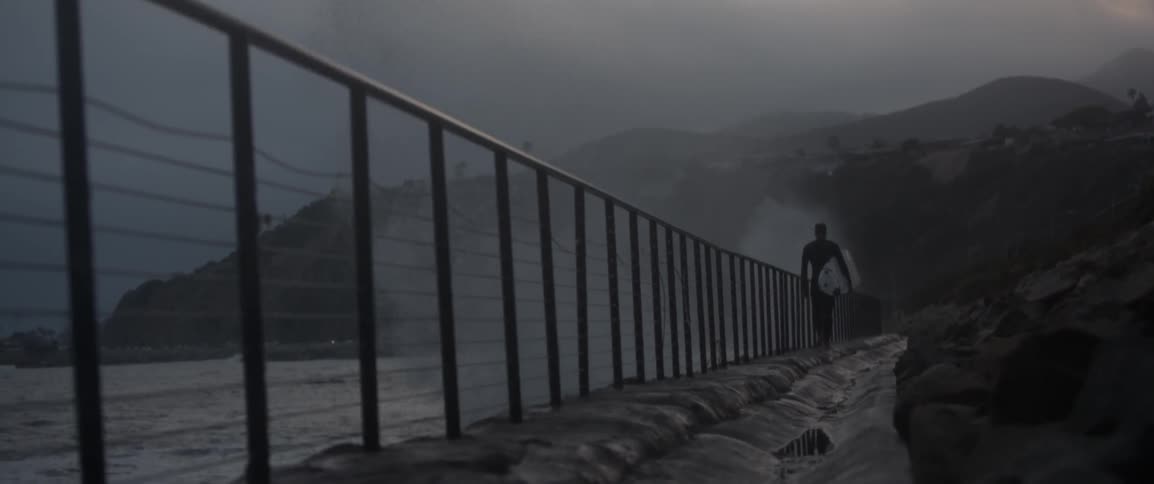 Ocean waves crash into a barrier and splash as a surfer walks by