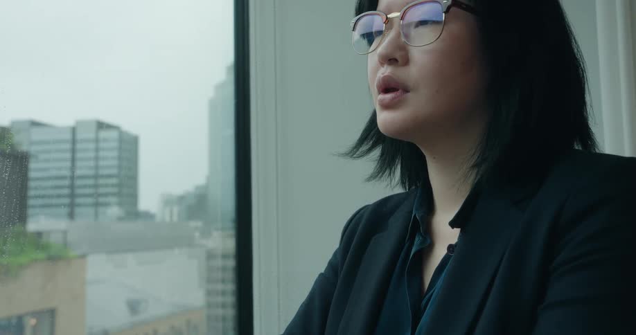 The video shows a closeup of a person with short black hair and glasses wearing a business blazer looking contemplatively out a highrise window the view outside is of a dense rainy city with tall buildings giving the scene a reflective or perhaps s