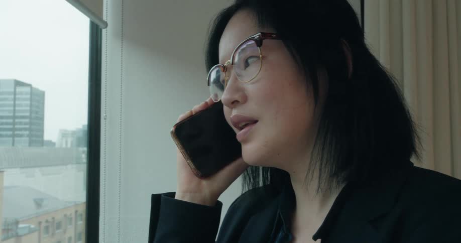 A young asian woman wearing a black blazer suit stands beside a glass wall inside an office building at midday making a phone call