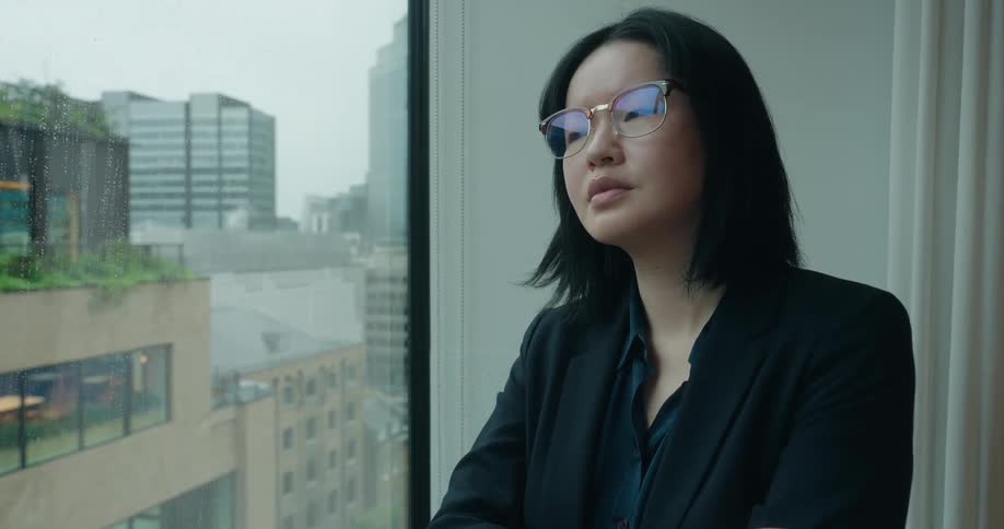 A young asian woman wearing a black suit and glasses stands by a glass window gazing at the buildings outside