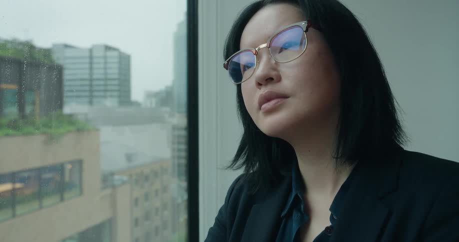 An asian woman looks out the window of an office building