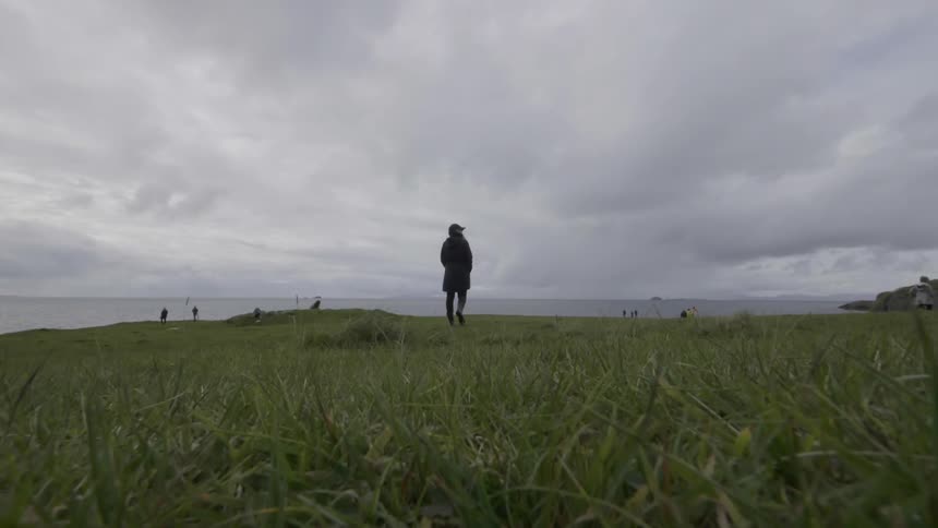 A woman walks on a grassy field overlooking the ocean