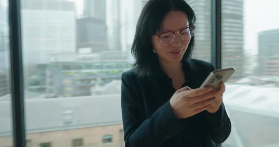 A smiling woman in a dark blazer and glasses stands by a large window looking down and typing on her smartphone in a modern high rise office