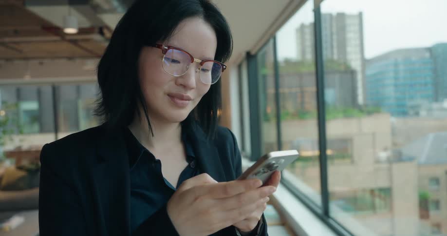 A professionally dressed woman with dark hair and glasses stands by a large window in a modern office looking down and smiling slightly while she interacts with a smartphone held in both hands with city visible outside the glass