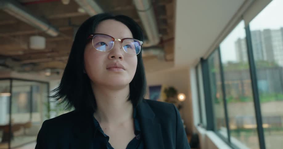 Young asian with glasses woman wearing black formal clothes looking out of a window over a metropolitan area indoors during day time