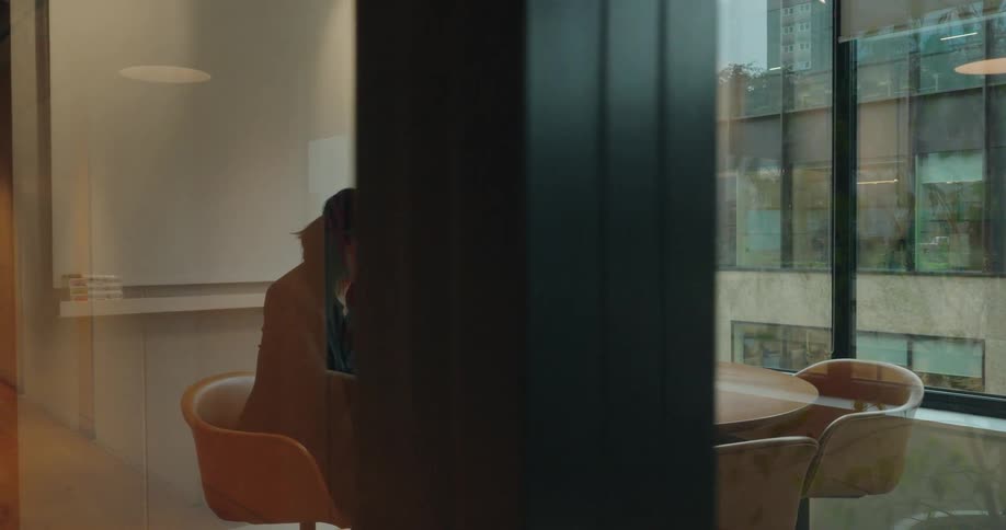 A woman dressed in dark clothing sits at a round wooden meeting table inside a welllit office space with large exterior windows focused on typing on a silver laptop computer