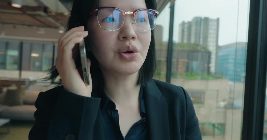 A professional woman in a black blazer and glasses talks seriously on a smartphone inside a modern office with large windows overlooking a city