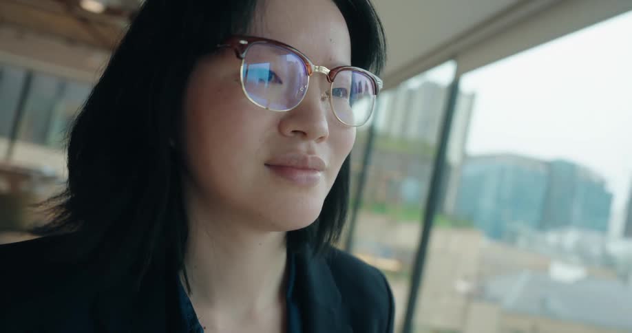 Woman in professional attire checks smartphone near window with urban view