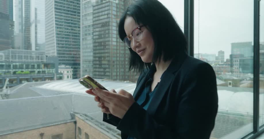 A young asian woman wearing a suit and glasses stands beside a window inside an office building smiling as she types on her mobile device with the city skyline visible through the glass