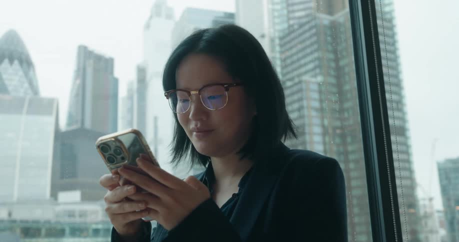 This video clip features a closeup of a woman indoors wearing glasses and a dark blazer looking at her smartphoneshe is standing in front of a large window that offers a view of a modern city skyline with several glass skyscrapers in the background