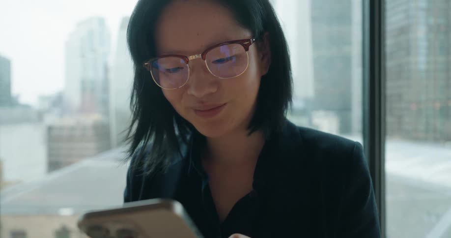 A woman is standing in front of a large window typing on her cellphone