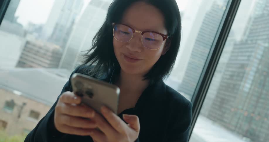 A young asian woman wearing a black suit and glasses stands beside a flower plant by a glass window at midday smiling as she types on her mobile phone with the city skyline visible in the background