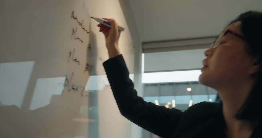 A young asian woman in a black suit and glasses writes on a whiteboard with a marker inside a conference room