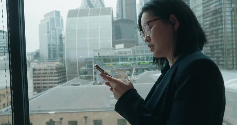 An asian woman texts on her phone while standing next to a window