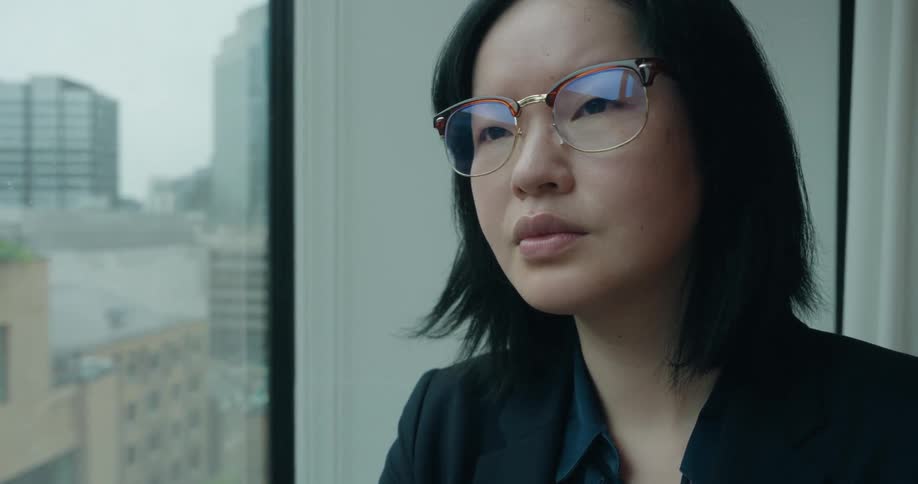 An asian woman in glasses stares out an office building window