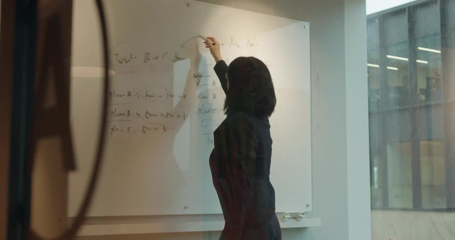 A woman in a dark blazer and glasses writes notes on a large clear glass whiteboard in a modern office meeting room